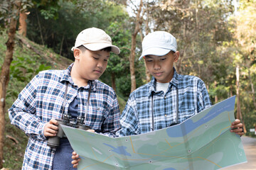 Asian young teen boys hold national park map, reading details of birdwatching before using their binoculars to watch the bird which sitting on brunches and flying in the sky, summer vacation concept.