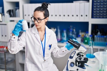 Young caucasian woman scientist using microscope holding pills bottle at laboratory