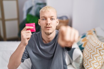 Young caucasian man holding condom sitting on bed pointing with finger to the camera and to you, confident gesture looking serious