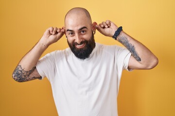 Young hispanic man with beard and tattoos standing over yellow background smiling pulling ears with...