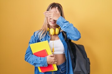 Young blonde woman wearing student backpack and holding books smiling and laughing with hand on face covering eyes for surprise. blind concept.