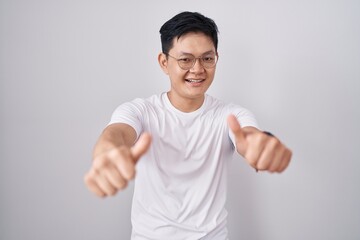 Fototapeta premium Young asian man standing over white background approving doing positive gesture with hand, thumbs up smiling and happy for success. winner gesture.