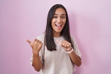 Young hispanic woman standing over pink background pointing to the back behind with hand and thumbs...