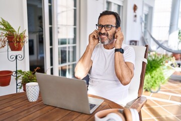 Middle age man using computer laptop at home covering ears with fingers with annoyed expression for the noise of loud music. deaf concept.