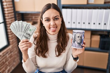 Young hispanic woman working at small business ecommerce holding money and piggy bank sticking tongue out happy with funny expression.