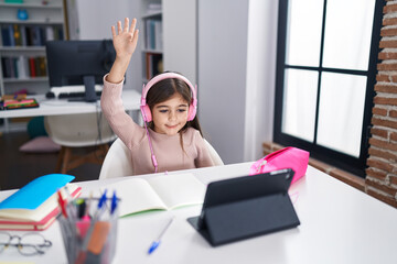 Adorable hispanic girl student using laptop and headphones with hand raised up at classroom