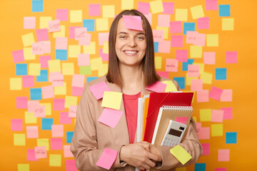 Indoor shot of smiling satisfied attractive Caucasian woman wearing beige jacket posing against yellow wall with colorful memo cards, holding paper folder, looking at camera, being in university.