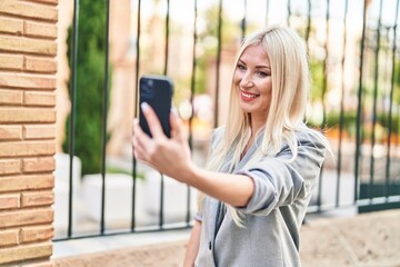 Young blonde woman smiling confident making selfie by the smartphone at street