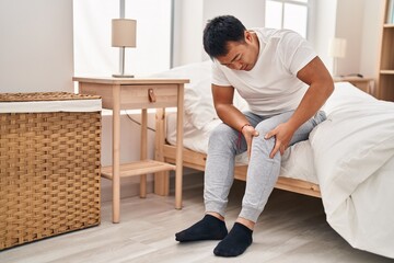 Young chinese man suffering for knee injury sitting on bed at bedroom