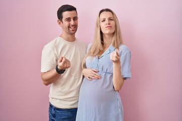 Young couple expecting a baby standing over pink background doing money gesture with hands, asking for salary payment, millionaire business