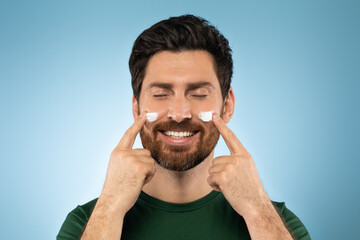 Happy bearded handsome man applying face cream on cheeks with closed eyes, posing over blue studio background