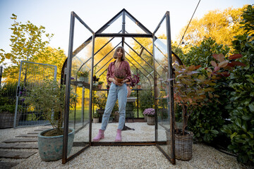 Full length portrait of a young cheerful woman florist or gardener standing near a beautiful vintage glasshouse for growing plants. Wide angle view