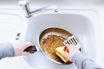woman's hand washes burnt greasy frying pan with kitchen washcloth in sink
