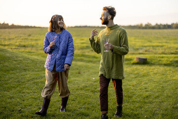 Stylish man and woman dressed warmly have conversation while walk together with wine on green lawn during a sunset. Couple spend leisure autumn time on nature