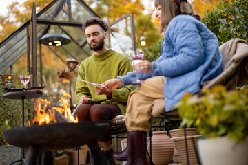 Young stylish couple grilling food and warming up while sitting together by the fire, spending autumn evening time at cozy atmosphere in garden