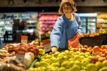 Young woman chooses apples to buy and puts them into mesh bag at fruit department of a supermarket. Concept of consumerism of healthy food and use of reusable bags