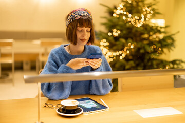 Fototapeta na wymiar Young adult woman works on a digital tablet and phone while sitting at modern coffee shop with Christmas tree on background. Concept of remote creative work online