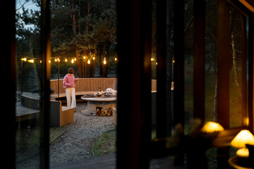Naklejka na ściany i meble Beautiful lounge area with round bench and bonfire illuminated with garlands near house in pine forest, view from the house