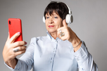 Woman 70-75 years old, wearing headphones and a shirt, talking using a smartphone via video link. Freelancer concept, female blogger, business