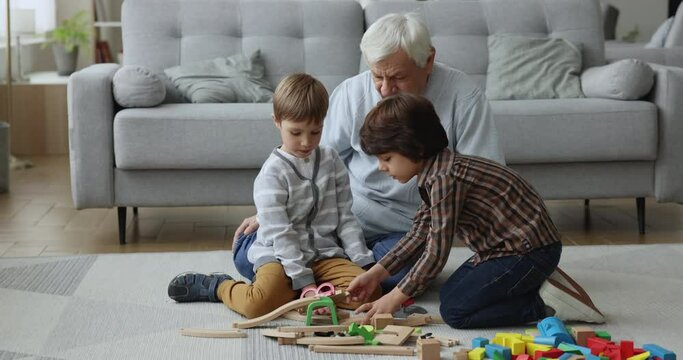 Two boys great-grandchildren play wooden railroad with great-grandfather, settled down on carpet in cozy living room enjoy game and friendly communication. Multi generational family leisure at home
