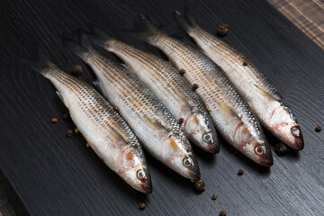 Five fresh silver mullet lies on a black board, surrounded by spices, close-up