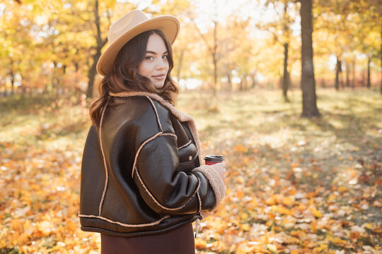 Cheerful young woman wearing in hat relaxing at park with yellow trees in background. Smiling girl enjoying warm sunny weather in autumn season.