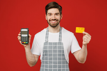 Young male housewife housekeeper chef cook baker man wear grey apron holding wireless modern bank payment terminal to process acquire credit card isolated on plain red background Cooking food concept.