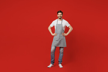 Fototapeta na wymiar Full body young happy housewife positive bachelor male housekeeper chef cook baker man wear grey apron stand akimbo arms on waist isolated on plain red color background studio. Cooking food concept.