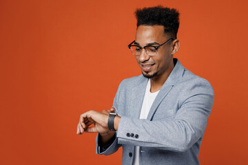 Young smiling happy employee business man corporate lawyer wear classic formal grey suit shirt glasses work in office use look at smart watch check time isolated on plain red orange background studio.