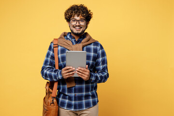 Young smiling teen Indian boy IT student wear casual clothes shirt glasses bag hold in hand digital tablet pc computer isolated on plain yellow color background High school university college concept