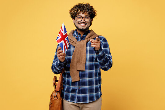 Young teen Indian boy IT student he wear casual clothes shirt glasses bag hold with British flag point finger camera on you isolated on plain yellow background. High school university college concept.
