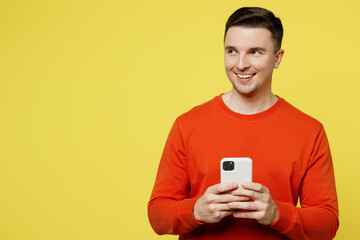 Young smiling cheerful happy man wear orange casual clothes hold in hand use mobile cell phone look aside on area isolated on plain yellow color background studio portrait. People lifestyle concept.