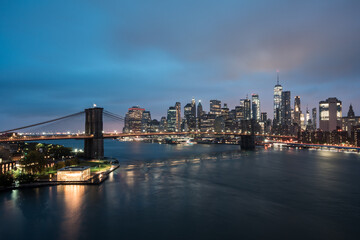Naklejka na ściany i meble Skyline von New York in der Dämmerung mit Brooklyn Bridge und Lichtern am Fluss.