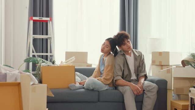Young Asian couple sit on sofa couch take a rest from organize moving carton box full of messy stuff in living room at new house. Husband and wife move new house, Apartment Rent And Ownership Concept.