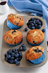 Freshly baked homemade blueberry muffins in a muffin tin with heap of fresh blueberries and sweet berry syrup on a white background. Selective focus, vertical.