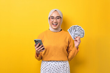 Beautiful cheerful Asian girl wearing hijab using mobile phone and holding money banknotes isolated on yellow background