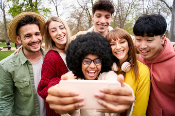 Obraz premium Young African girl taking a selfie portrait of her large group of friends hugging and smiling. Cheerful people looking phone in outdoor park. Concept of communication, community and friendship.