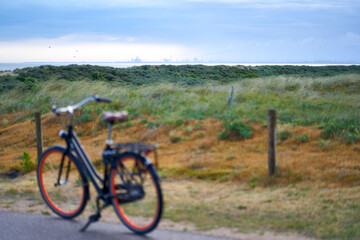 Fototapeta na wymiar Port building of Rotterdam on horizon at dawn. Ladies bicycle in depth blur in front of green dunes landscape. Netherlands, South Holland, Ouddorp.