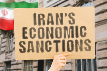 The phrase " Iran's Economic Sanctions " is on a banner in men's hands with blurred background. Manufacturing. Exportation. Import. Supply. Demand. Shortage. International. Diplomacy. Negotiation