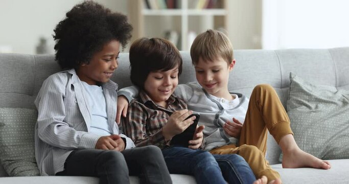 Three multi racial boys having fun at home using smartphone. Friends sit on sofa spend free time on internet, watch videos on-line, play games, use mobile application. Young gen and modern tech usage