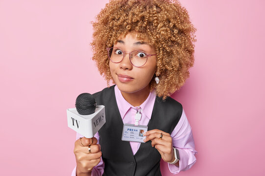 Professional female reporter holds microphone going to give speech to audience looks with wonder at camera dressed in formal clothes shows badge with personal information isolated over pink background