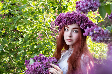 young girl with long red hair with a wreath of lilacs on her head in the garden in spring