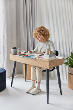 Indoor shot of curly female artist poses at table draws sketches or pictures dressed in casual domestic clothes poses against cozy home interior. People hobby pastime and creativity concept.