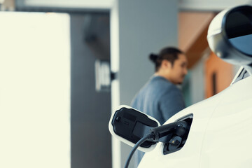 Focus electric car charging at home charging station with blurred progressive man walking in the...