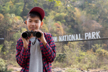 Asian teen boy holds binocular and standing by sling bridge in local national park before watching...