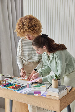 Two female designers work together as team draw interior design for future apartment choose color of walls from palette stands near wooden table with crayons and papers involved in creative process