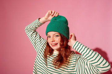 Happy smiling freckled redhead teenager girl with dental retainer, wearing trendy green beanie hat, striped long sleeve shirt, posing on pink background. Copy, empty space for text - obrazy, fototapety, plakaty