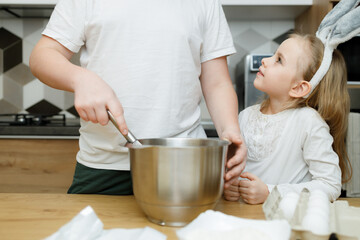 Siblings stand at kitchen table. Little girl in bunny ears looking at her big brother whisking dough in modern kitchen. Easter concept