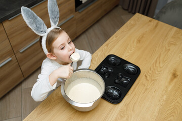 Little girl in bunny ears eating, licking sweet dough for muffins or cake with spoon from bowl in modern kitchen