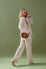 Fashionable confident woman wearing elegant white suit with blazer, wide leg trousers, trendy sunglasses, brown suede shoulder bag, posing on green background. Full-length studio fashion portrait - 565953075
